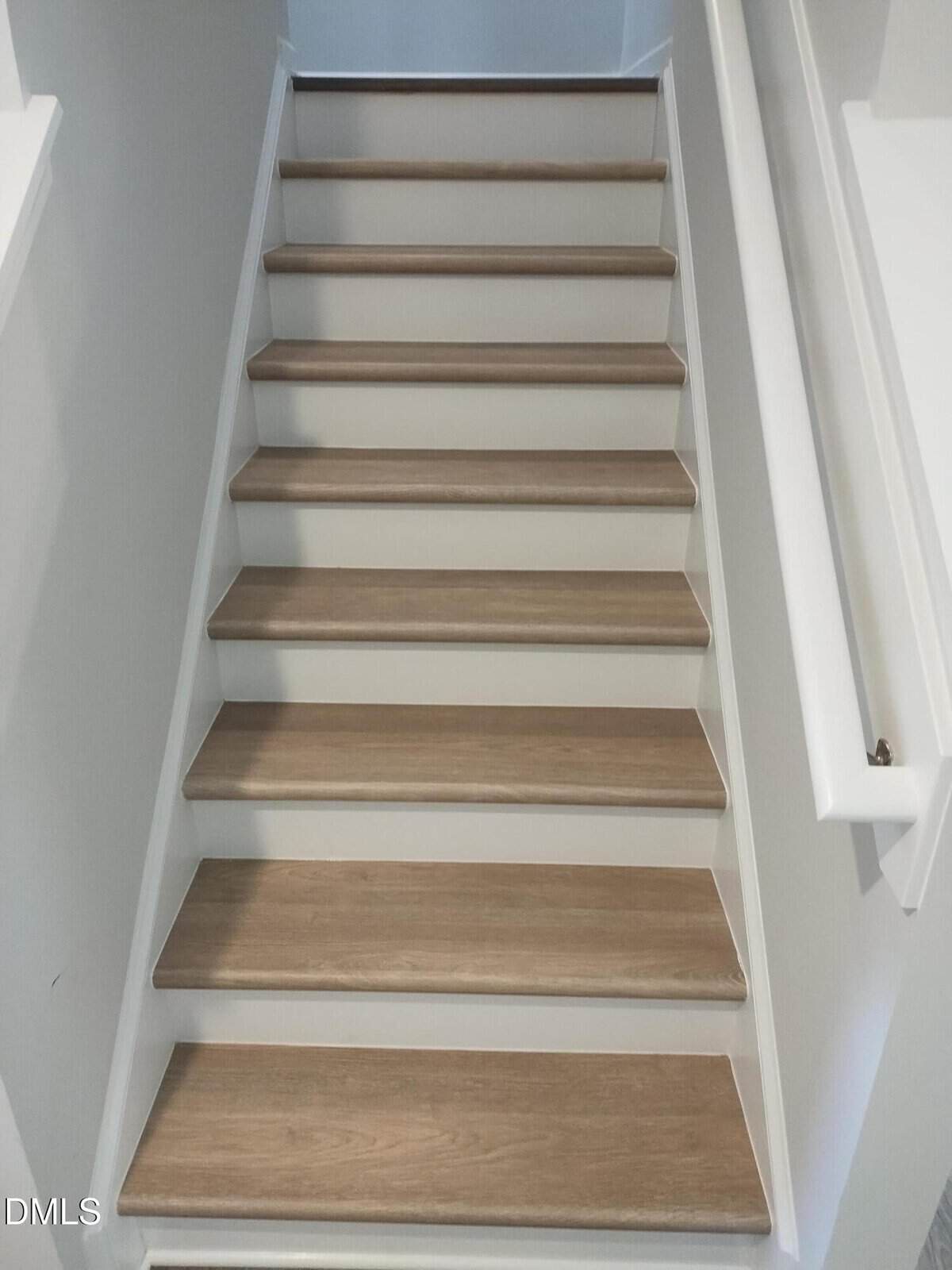 A straight staircase with light wood steps and white risers and walls, viewed from the bottom looking up. There’s a white handrail on the right side.