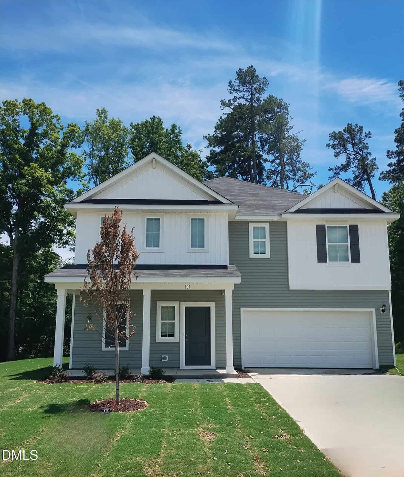 Modern two-story house at 101 Airdale Drive, Oxford, with gray siding, white trim, and a double garage. A small tree accents the front yard with green grass and a concrete driveway. Tall trees line the background under a blue sky.