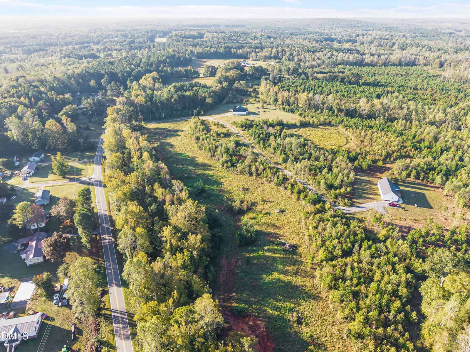 Aerial view of a rural landscape featuring a winding road, scattered houses, dense green trees, grassy fields, and a large barn-like building on the right side of the image.