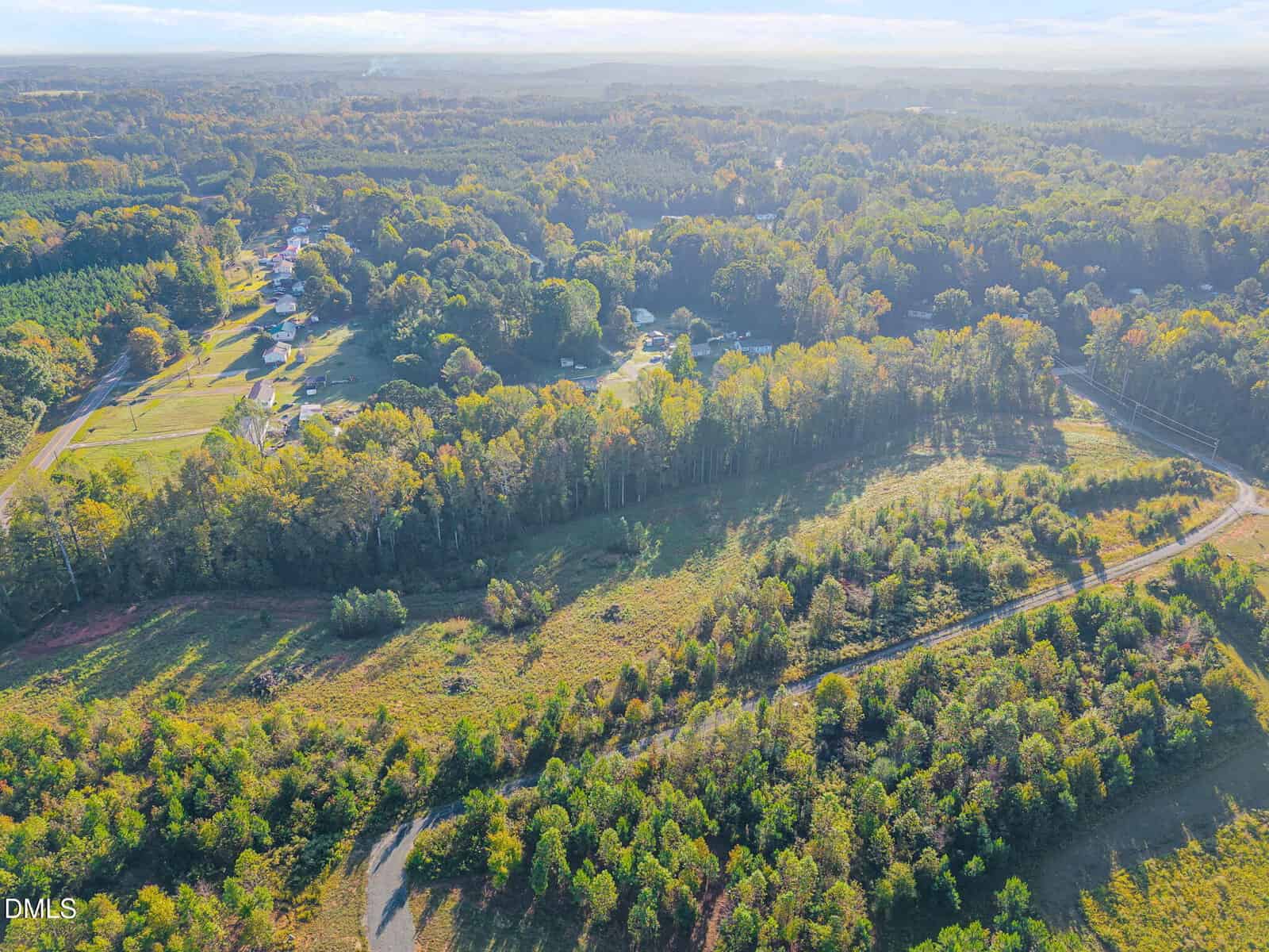 Aerial view of a rural landscape with a mix of open fields, wooded areas, a winding road, and scattered houses nestled among trees under a partly cloudy sky.