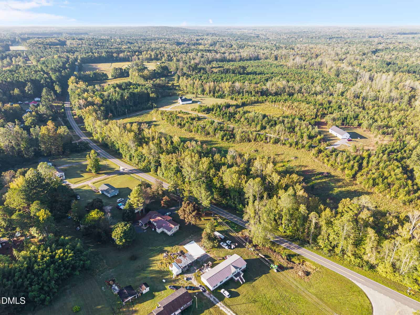 Aerial view of a rural landscape with houses, roads, open fields, and dense green trees under a blue sky. Several properties are visible, surrounded by forest and meadows.