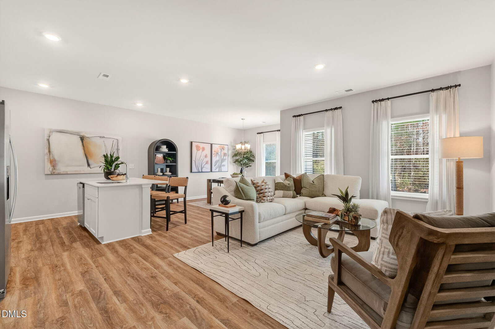Modern open-concept living room and kitchen with wood flooring, white cabinets, a beige sofa, armchairs, large windows with white curtains, and decorative plants. Neutral tones and contemporary decor throughout.