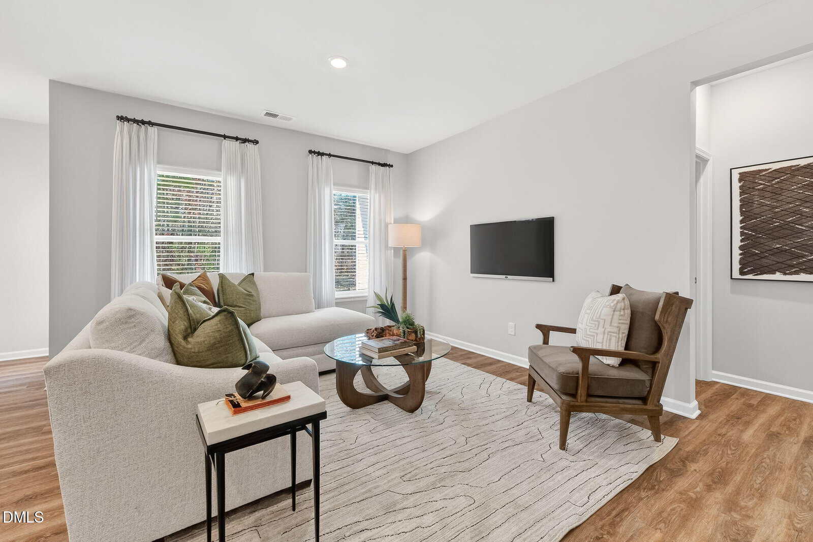 Bright living room with a white sectional sofa, green and beige pillows, wooden armchair, glass coffee table, TV on wall, floor lamp, large windows with white curtains, and a textured rug on wooden floors.