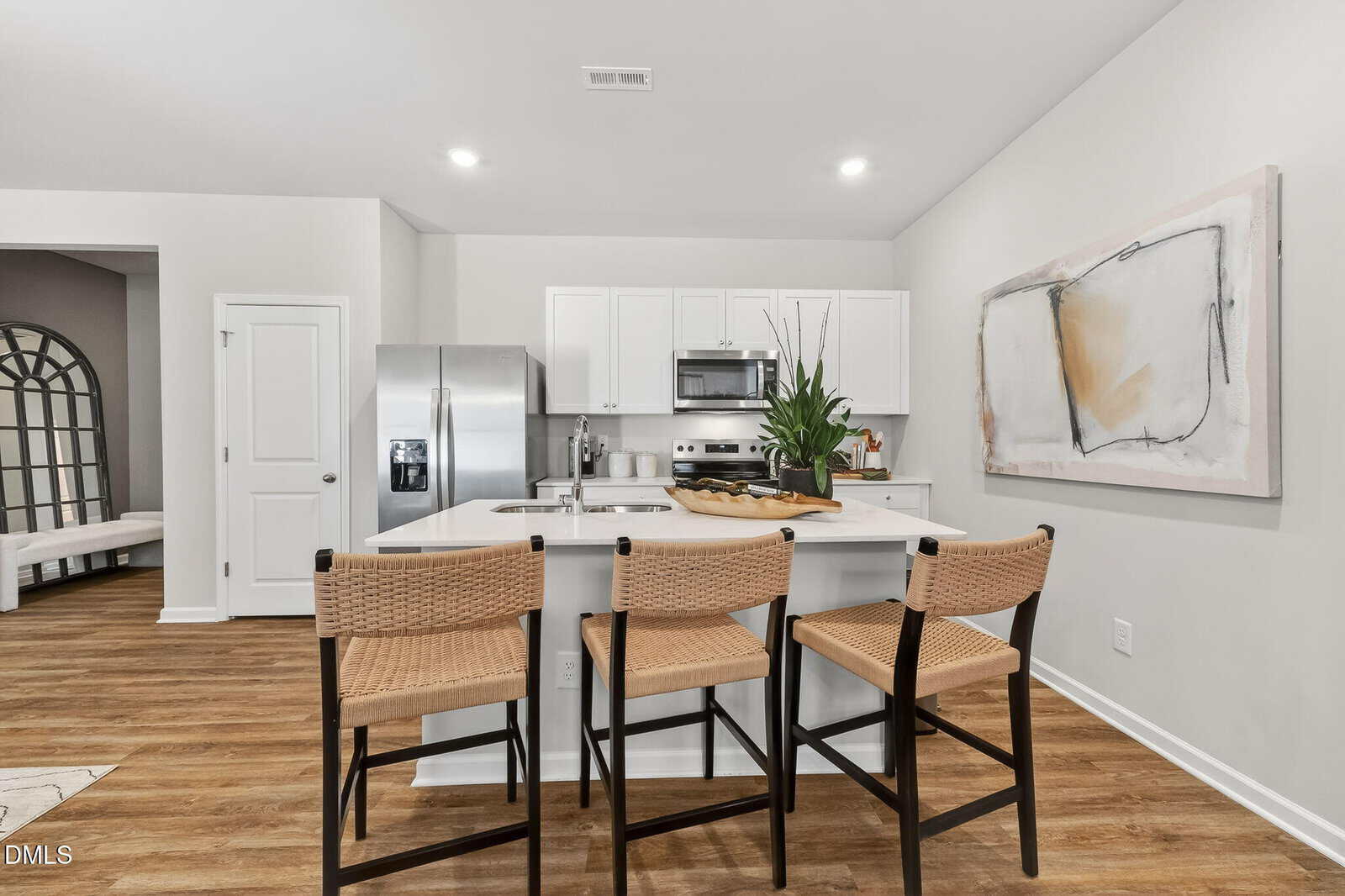 Modern kitchen with white cabinets, stainless steel appliances, a central island with three woven barstools, a plant centerpiece, abstract wall art, and wood flooring. A doorway leads to another room.