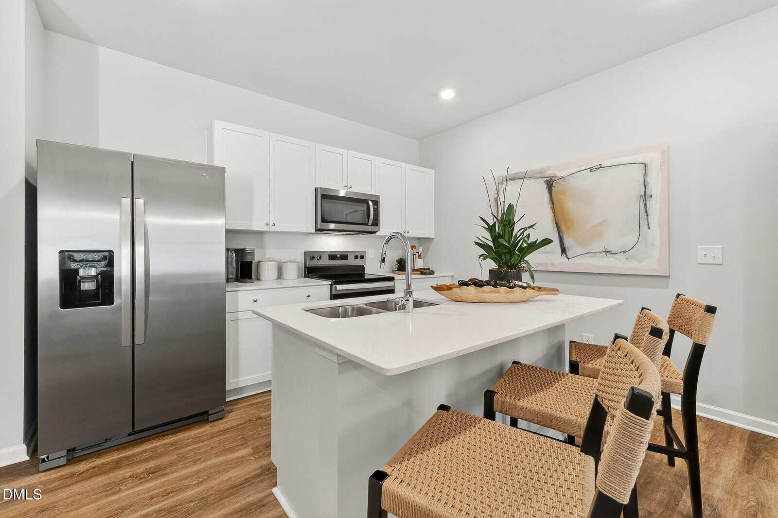 Modern kitchen with white cabinets, stainless steel appliances, island with sink, three woven chairs, a plant centerpiece, wooden floors, and abstract art on the wall.
