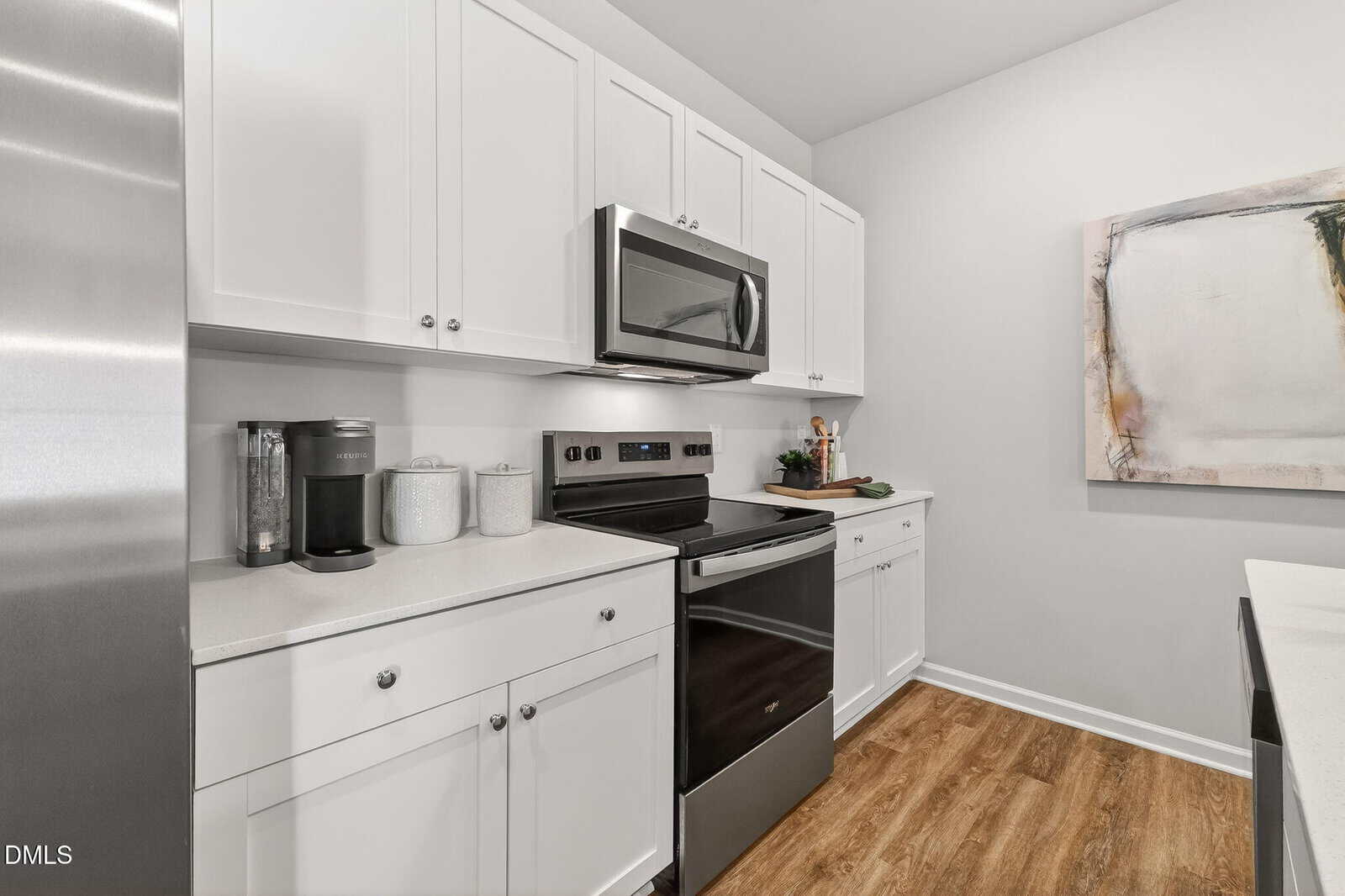 Modern kitchen with white cabinets, stainless steel appliances, a coffee maker, and a large abstract painting on the wall above wood-look flooring. Counters are clean and minimal.