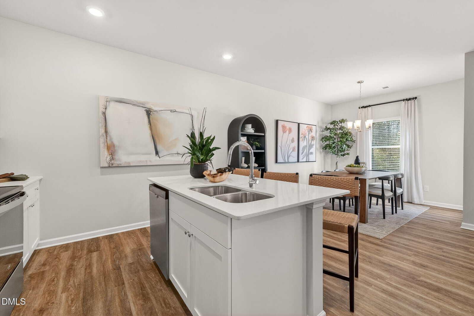 Modern kitchen with a white island, sink, and bar stools, open to a dining area with a table, chairs, large windows, and botanical artwork on the walls. Wood flooring and neutral tones create a bright, inviting space.