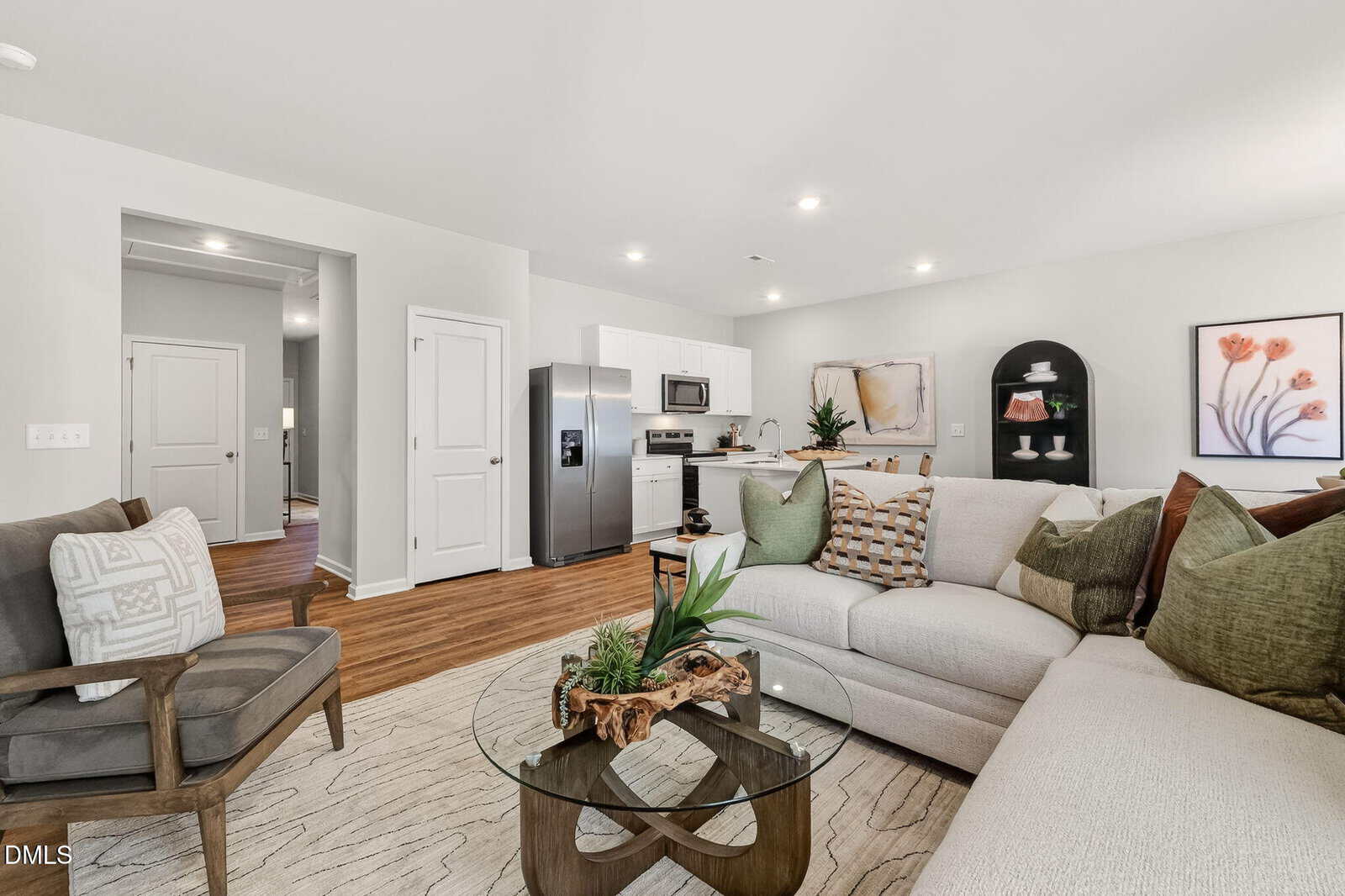 Modern living room with a sectional sofa, round glass coffee table, armchair, and open kitchen in the background at 103 Airdale Drive. Neutral decor with green accents, wall art, plants, and wooden floors create a cozy, inviting space infused with the timeless charm of Oxford living.