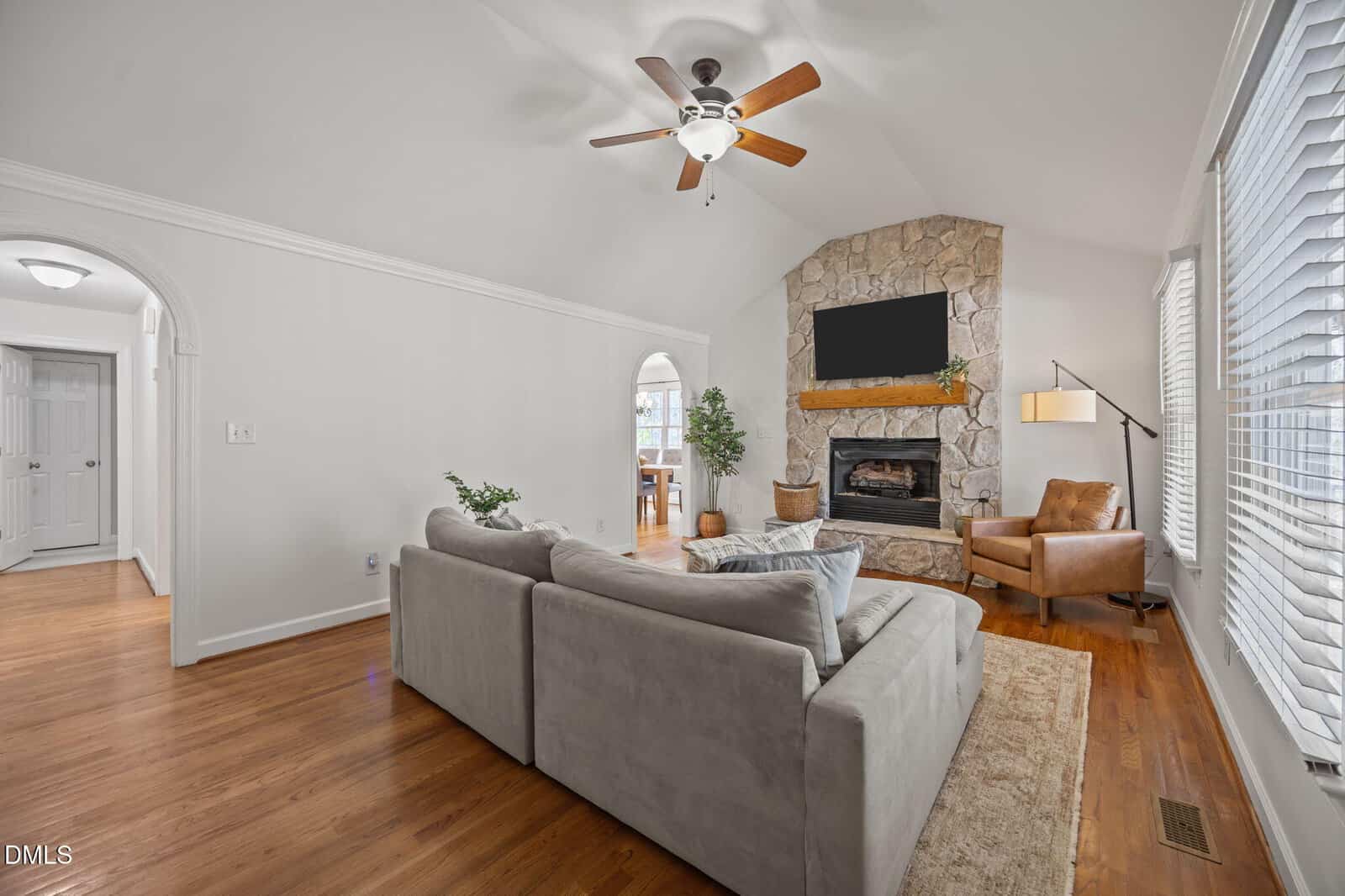 A cozy living room with a gray sectional sofa, stone fireplace, TV above mantel, leather armchair, floor lamp, large window with blinds, wood floors, and white walls. An arched doorway leads to a dining area.