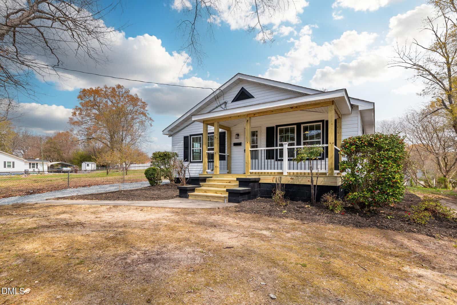 A small, white, single-story house with a covered front porch, black trim, and wooden steps. The yard is mostly bare with some bushes, and neighboring houses are visible in the background under a partly cloudy sky.