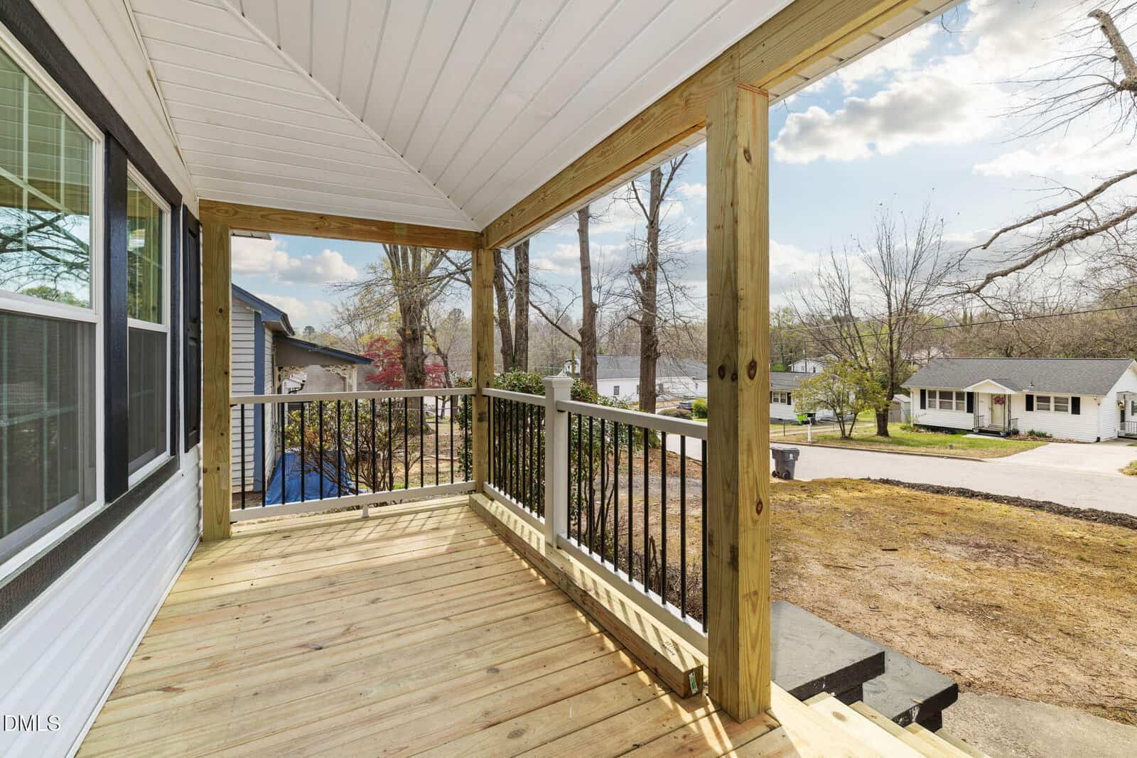 A wooden front porch with a white ceiling and railings featuring vertical black balusters, overlooking a residential street with houses, trees, and a partly cloudy sky in the background.