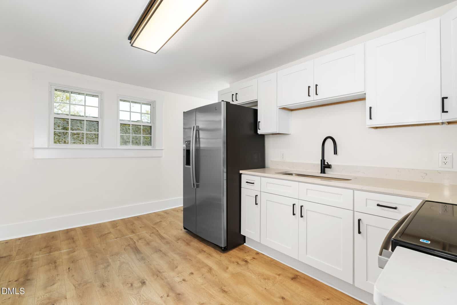Modern kitchen with white cabinets, black hardware, stainless steel refrigerator, stove, and dishwasher, light wood flooring, large window, and white walls.