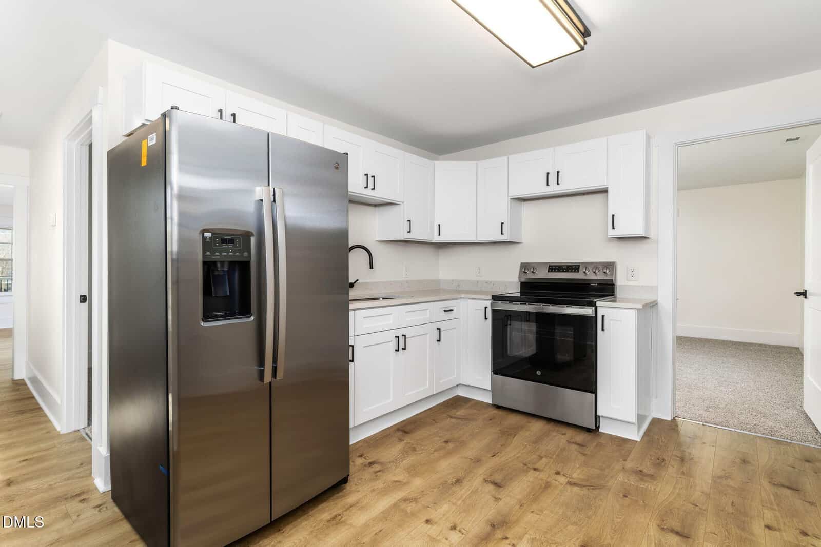 Modern kitchen with stainless steel refrigerator, oven, and white cabinets. The space features light wood flooring, under-cabinet lighting, and an open doorway leading to a carpeted room.