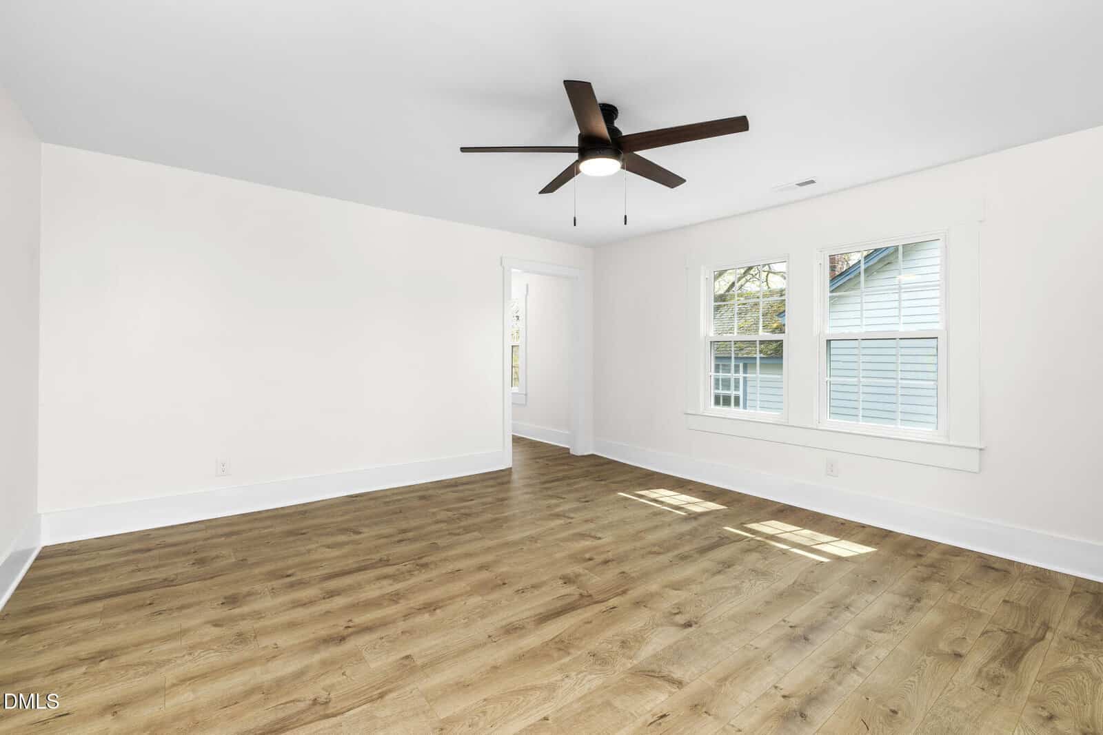 An empty room with white walls, light wood flooring, a ceiling fan with lights, and two windows showing the exterior of a neighboring house. Natural light brightens the space.