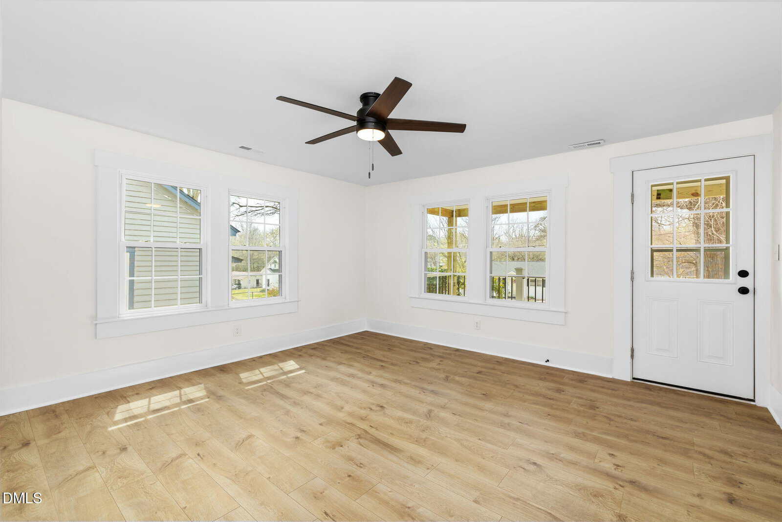 Bright, empty room with light wood flooring, white walls, large windows, a black ceiling fan, and a white door leading outside. Sunlight streams through the windows, giving the space an airy feel.