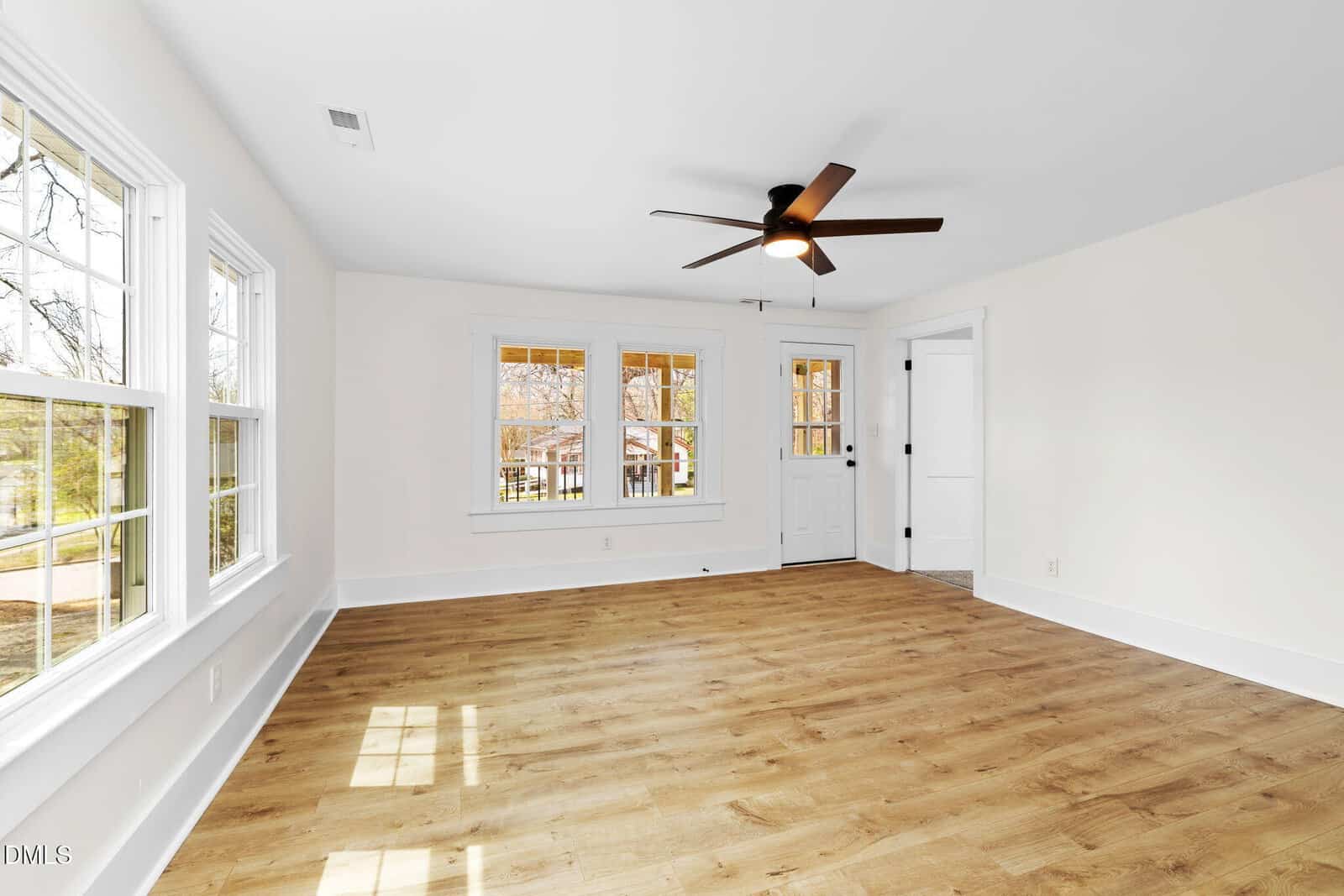 A bright, empty room with white walls, wood-style flooring, multiple windows, a white door with glass panes, and a ceiling fan with a light fixture. Natural light fills the space through the windows.