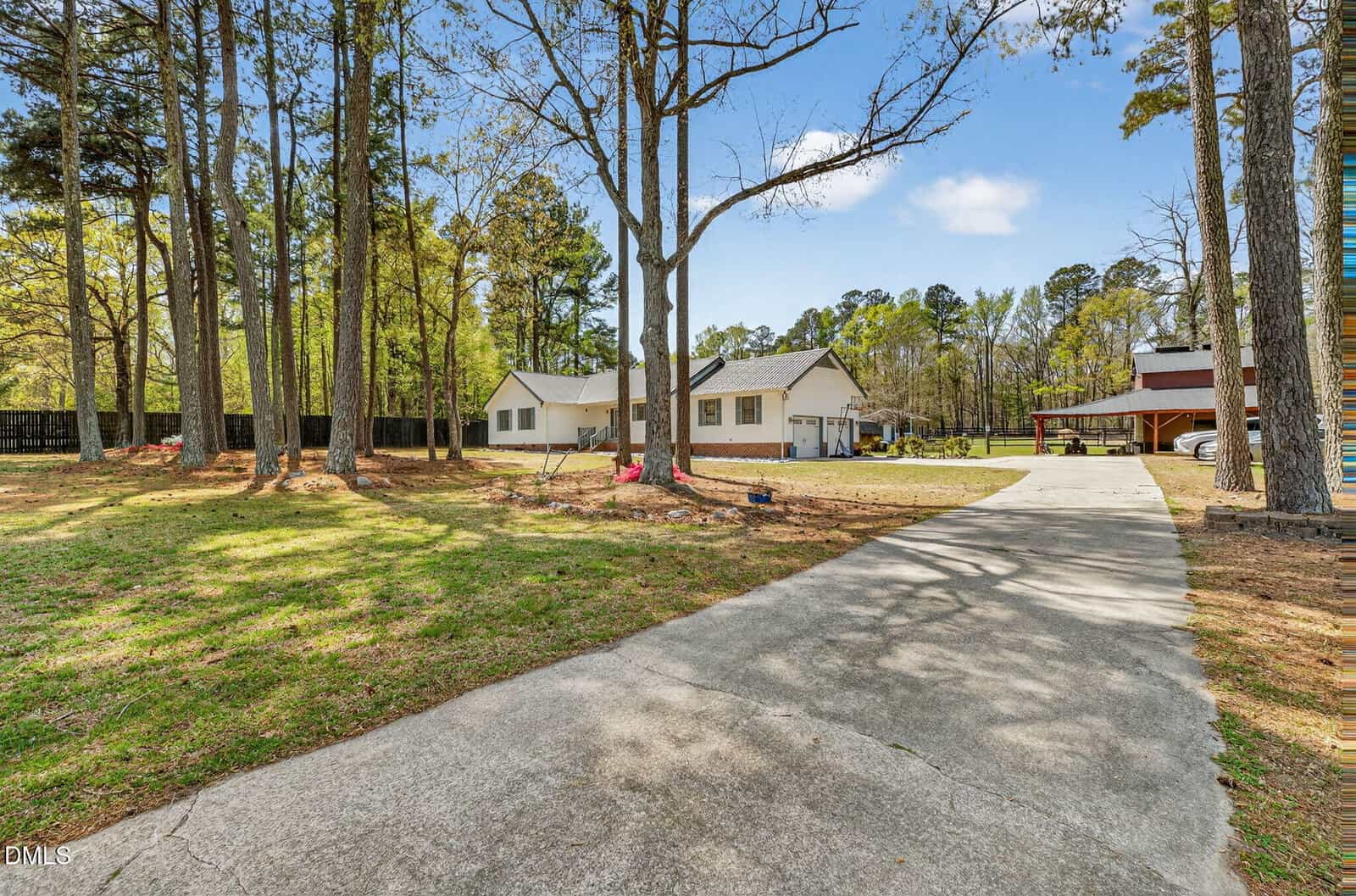 A concrete driveway leads to a white single-story house with a detached garage and a separate brown building. Tall trees and a grassy yard surround the property under a clear blue sky.