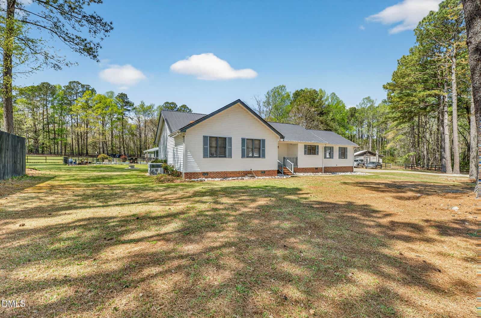 A single-story white house with a dark roof sits on a large grassy lot surrounded by trees under a clear blue sky. A fenced area and another structure are visible in the background.