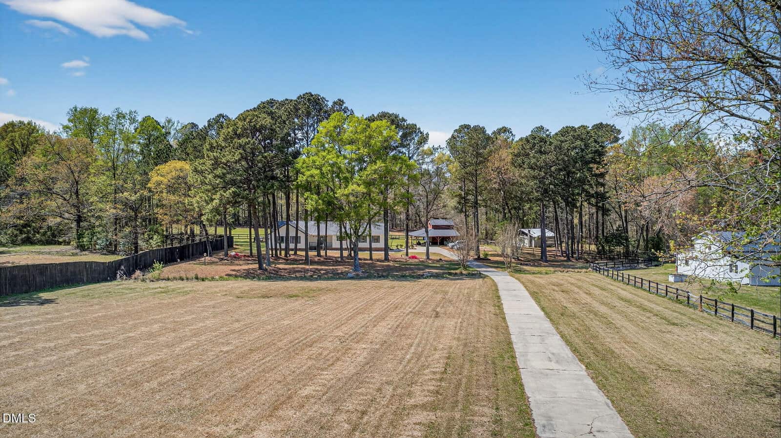 A long paved driveway leads through a large, grassy field toward several buildings and tall trees. The area is fenced, with houses and farm structures visible among the trees under a blue sky.