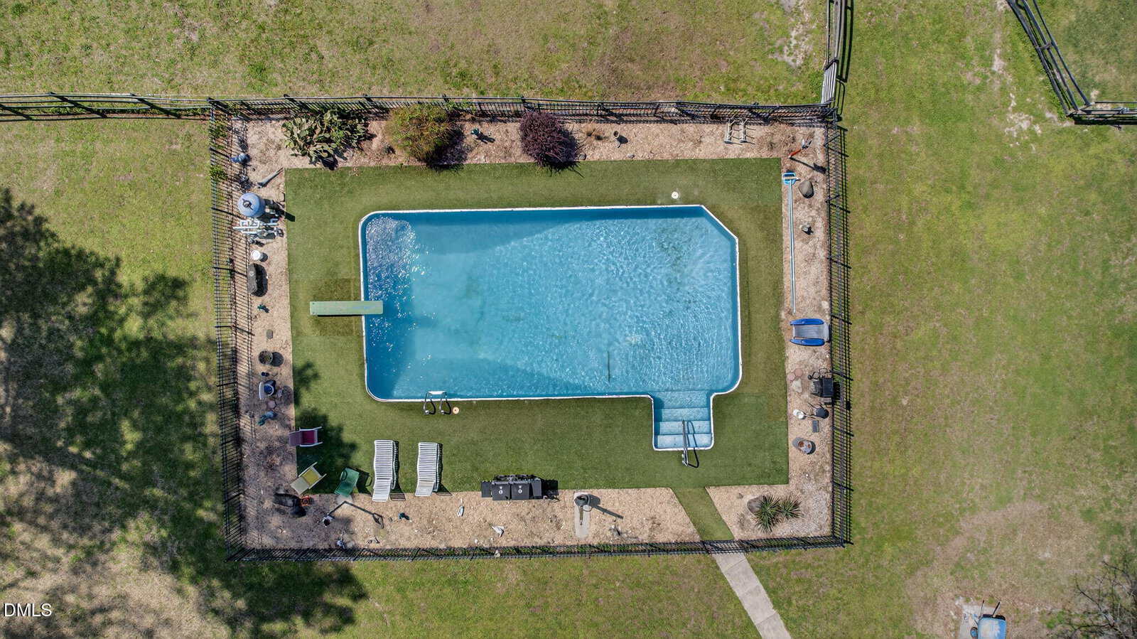 Aerial view of a rectangular swimming pool with a diving board, steps, and poolside lounge chairs, surrounded by grass and enclosed by a fence.