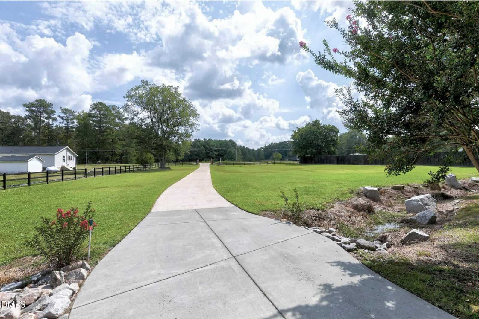 A long, curved concrete driveway leads through a large grassy yard with scattered trees, a white fence, a small white building on the left, rocks on the right, and a partly cloudy sky overhead.