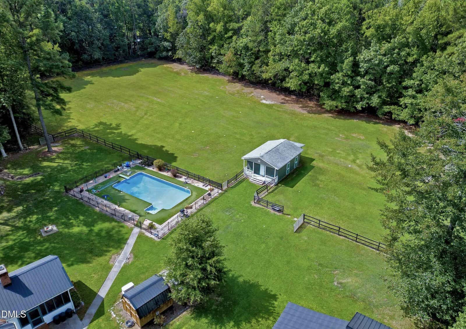 Aerial view of a large grassy yard with a small house, fenced-in swimming pool, and several trees lining the property. The area is bordered by dense woods.
