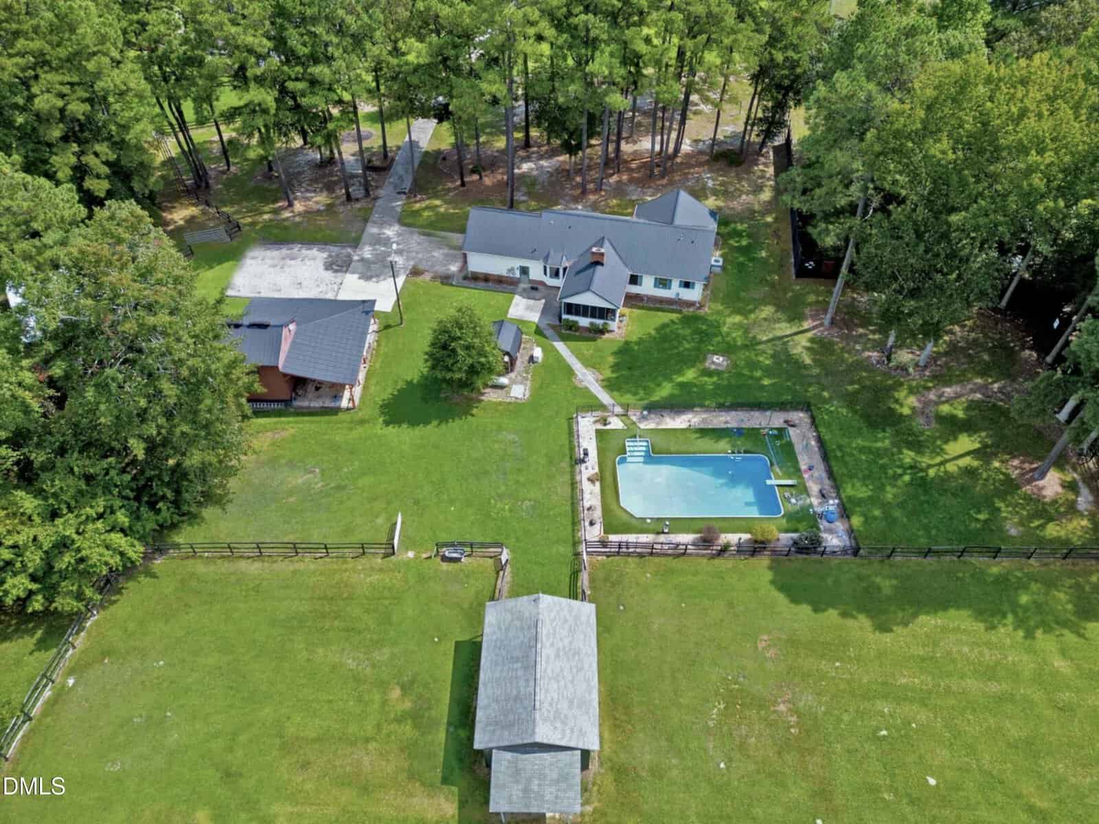 Aerial view of a property with multiple buildings, a driveway, a rectangular swimming pool with a fenced area, and large green lawn surrounded by trees.
