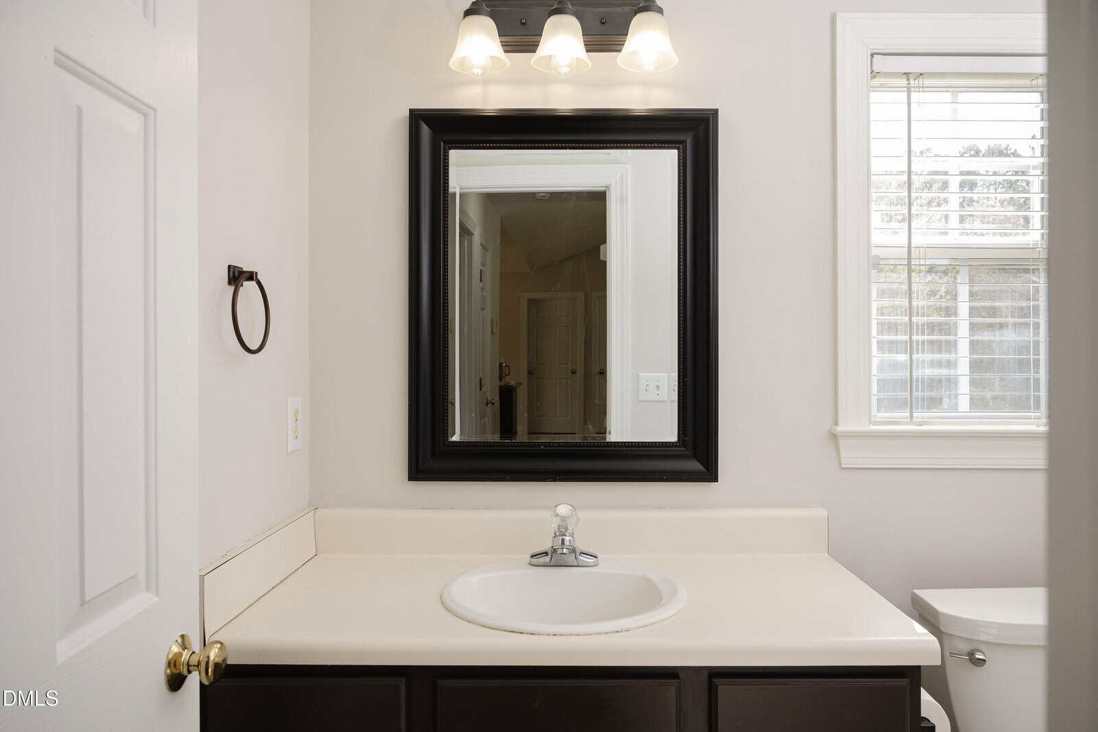 White bathroom vanity with single sink, black-framed mirror above, three-light fixture, towel ring on the left wall, and a window with white blinds on the right, partially showing the toilet.