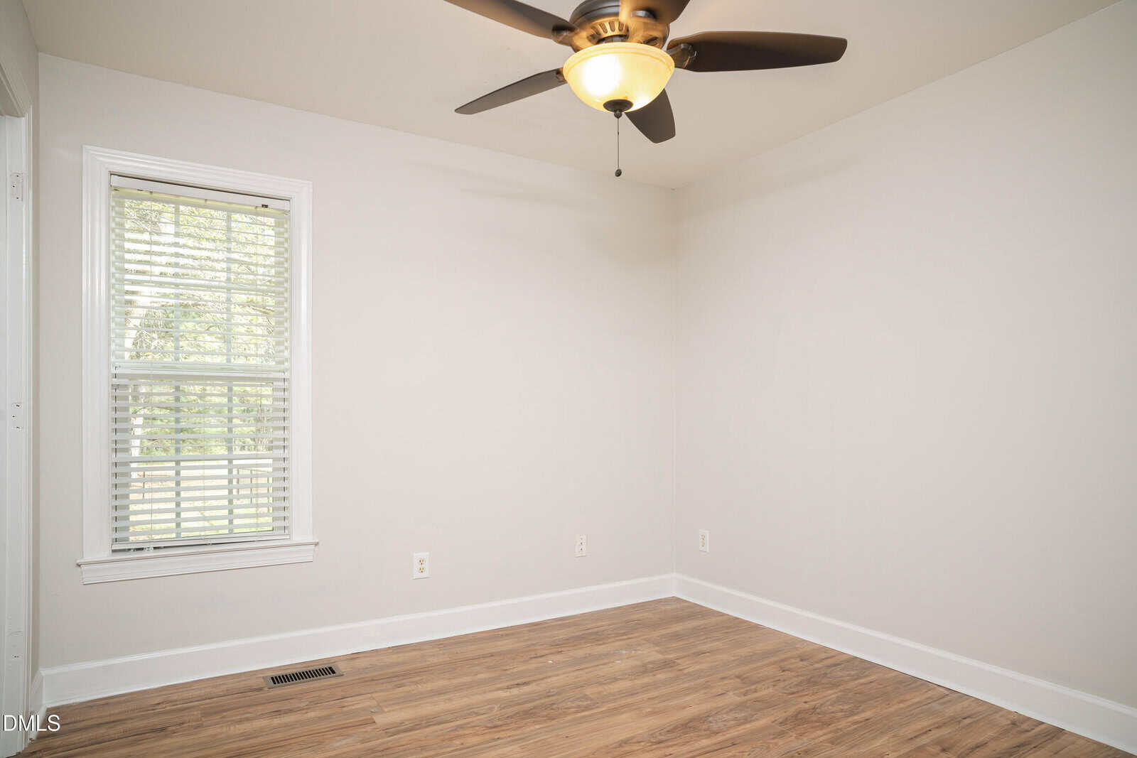 A small, empty room with light-colored walls, a window with white blinds, wood flooring, and a ceiling fan with a light.
