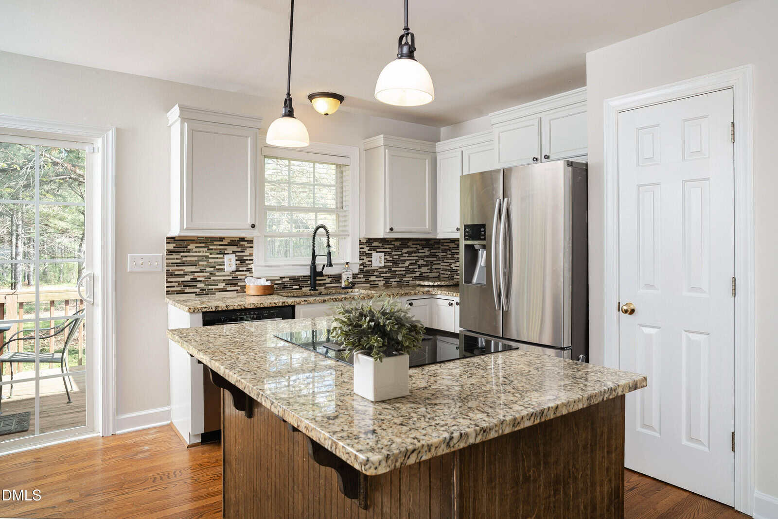 Modern kitchen with white cabinets, stainless steel appliances, granite countertop island with a potted plant, tile backsplash, pendant lights, wood floors, and a window overlooking an outdoor deck.