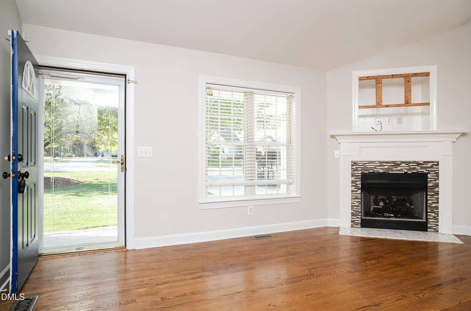 A bright room with hardwood floors, a blue front door open to the outside, large window with blinds, and a white fireplace with a decorative tile surround.
