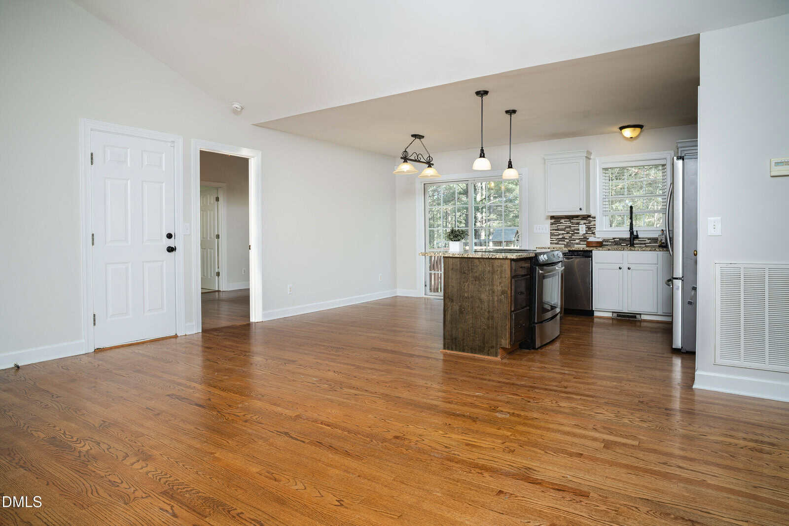 Open-concept room with hardwood floors, white walls, and a kitchen area featuring white cabinets, an island with a stove, pendant lights, and large windows letting in natural light. A white door is visible on the left.