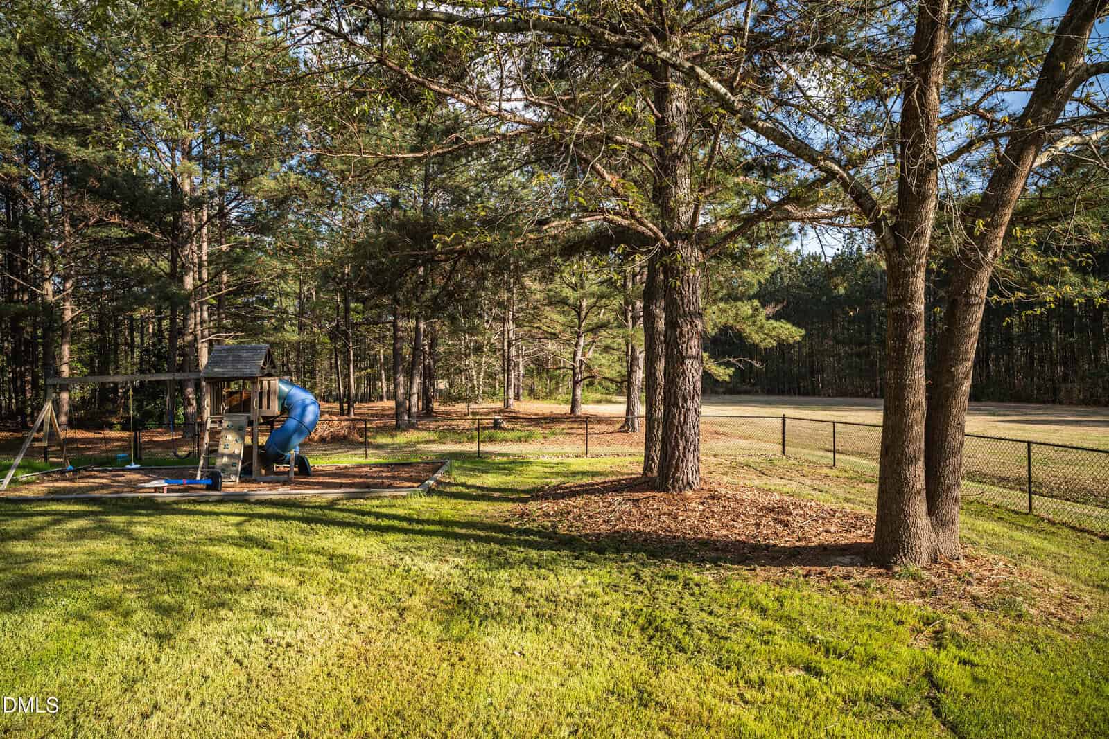 A person in blue coveralls works on a small structure in a grassy, tree-filled yard bordered by a wire fence, with scattered sunlight filtering through the tall pine trees.