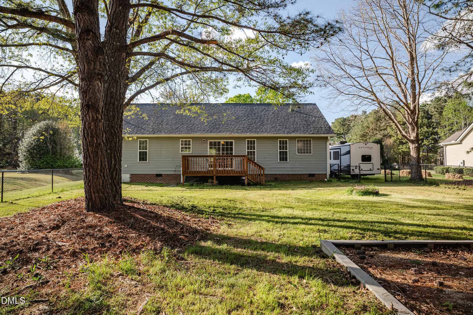 A single-story house with gray siding and a small wooden deck is shown from the backyard, surrounded by grass, a large tree, and a fenced area. An RV is parked on the right side near the house.