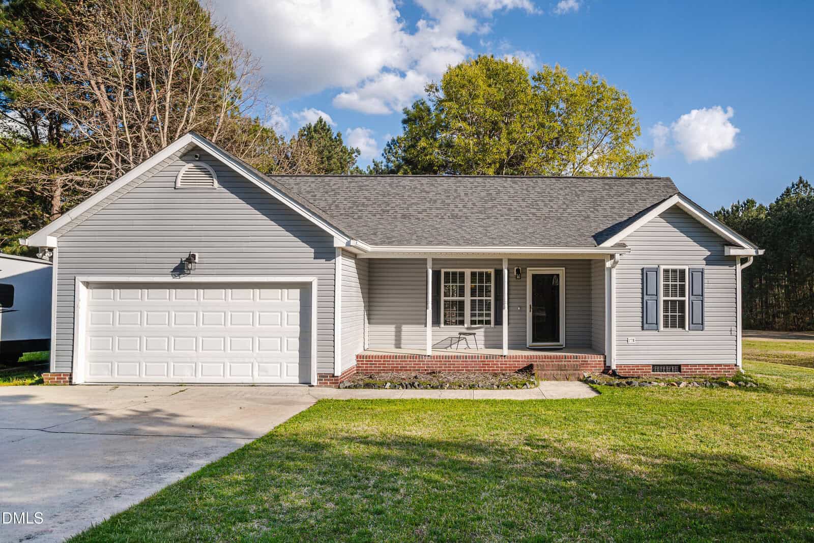 A single-story gray house with a two-car garage, white trim, and a covered front porch, set on a grassy lawn with trees in the background under a partly cloudy sky.