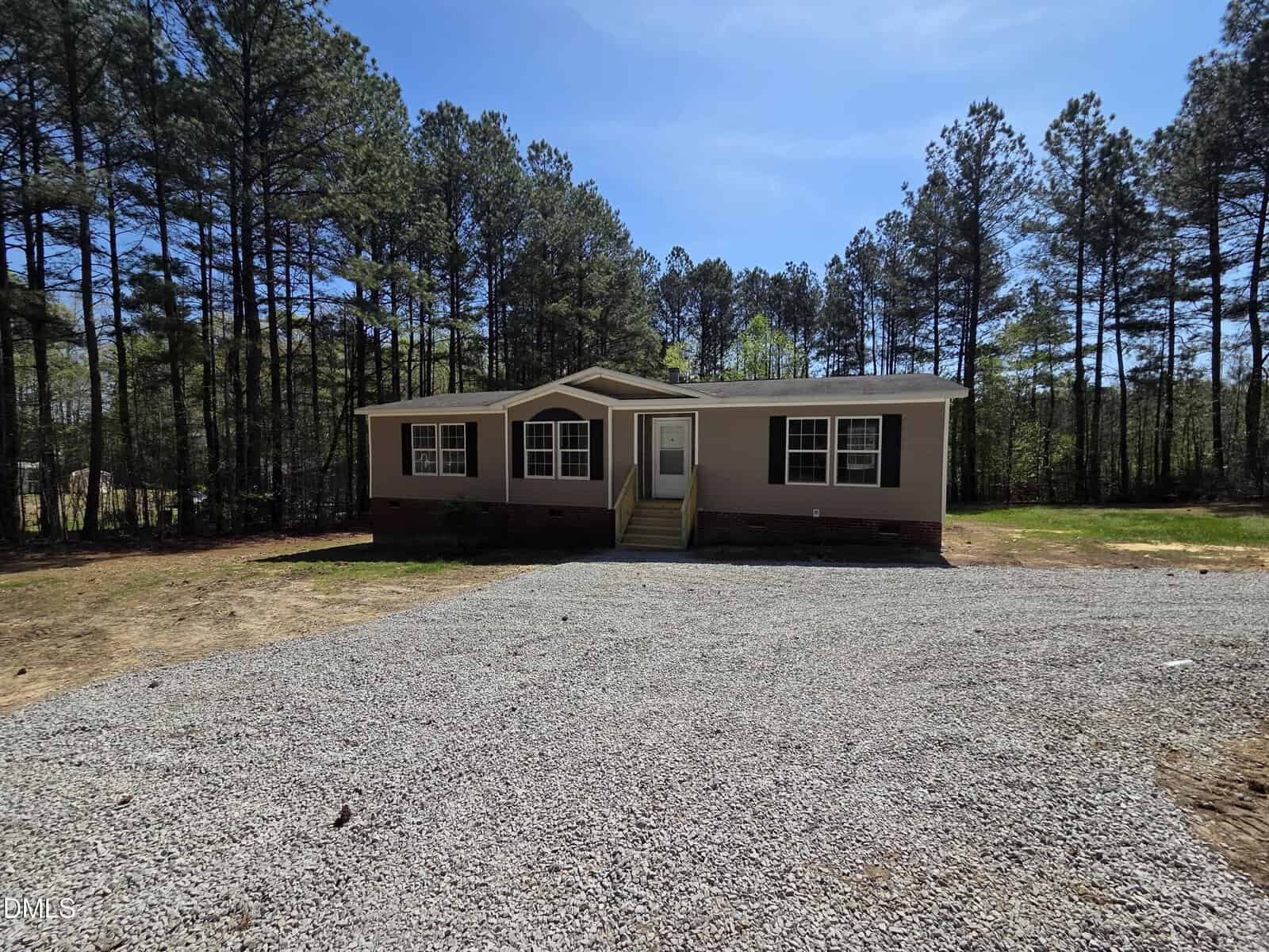 A single-story modular home with light-colored siding and a front porch, set on a gravel driveway and surrounded by tall pine trees under a clear blue sky.
