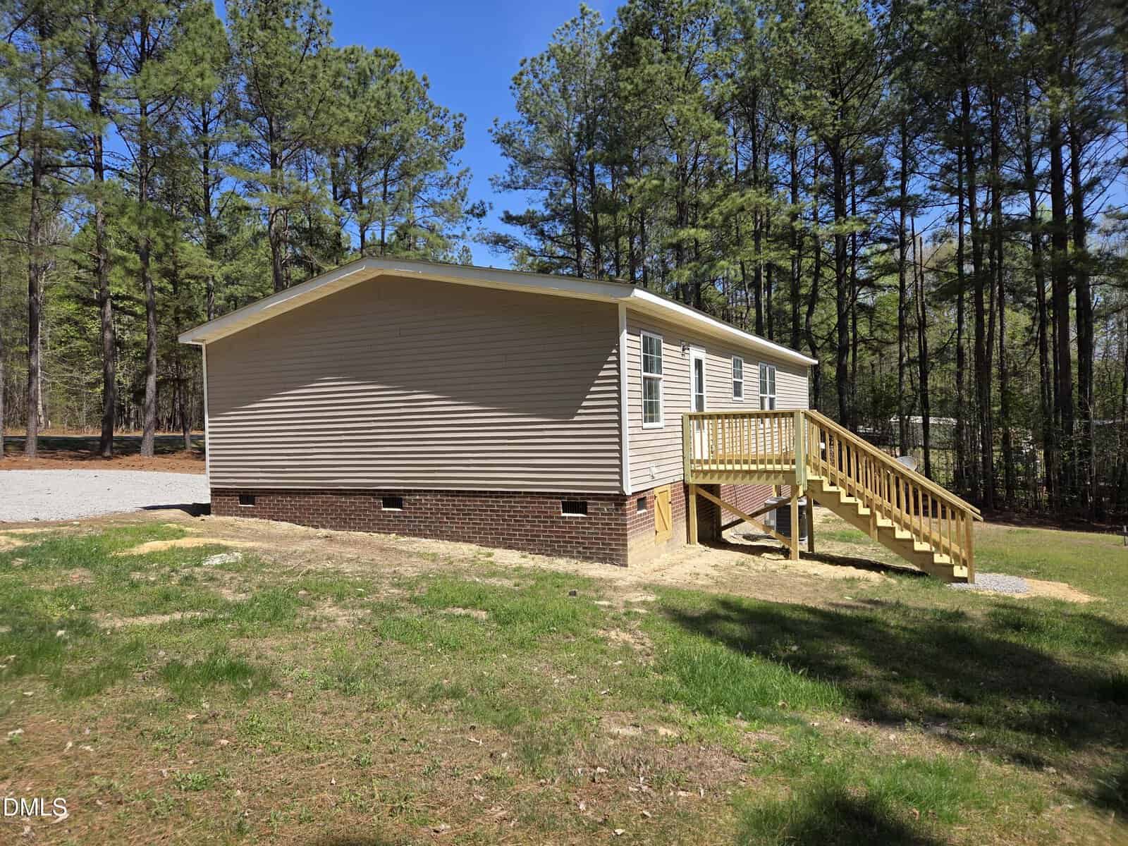 A single-story beige house with a brick foundation, four windows, and a wooden staircase leading to the front door, surrounded by grass and tall pine trees.