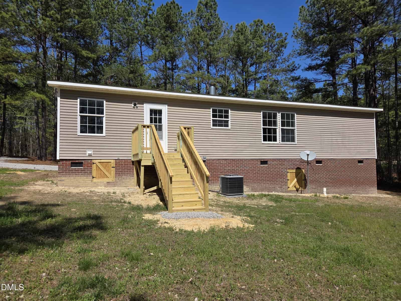 A single-story beige house with brick foundation, front steps leading to a white door, multiple windows, an air conditioning unit, satellite dish, and surrounded by trees under a clear blue sky.
