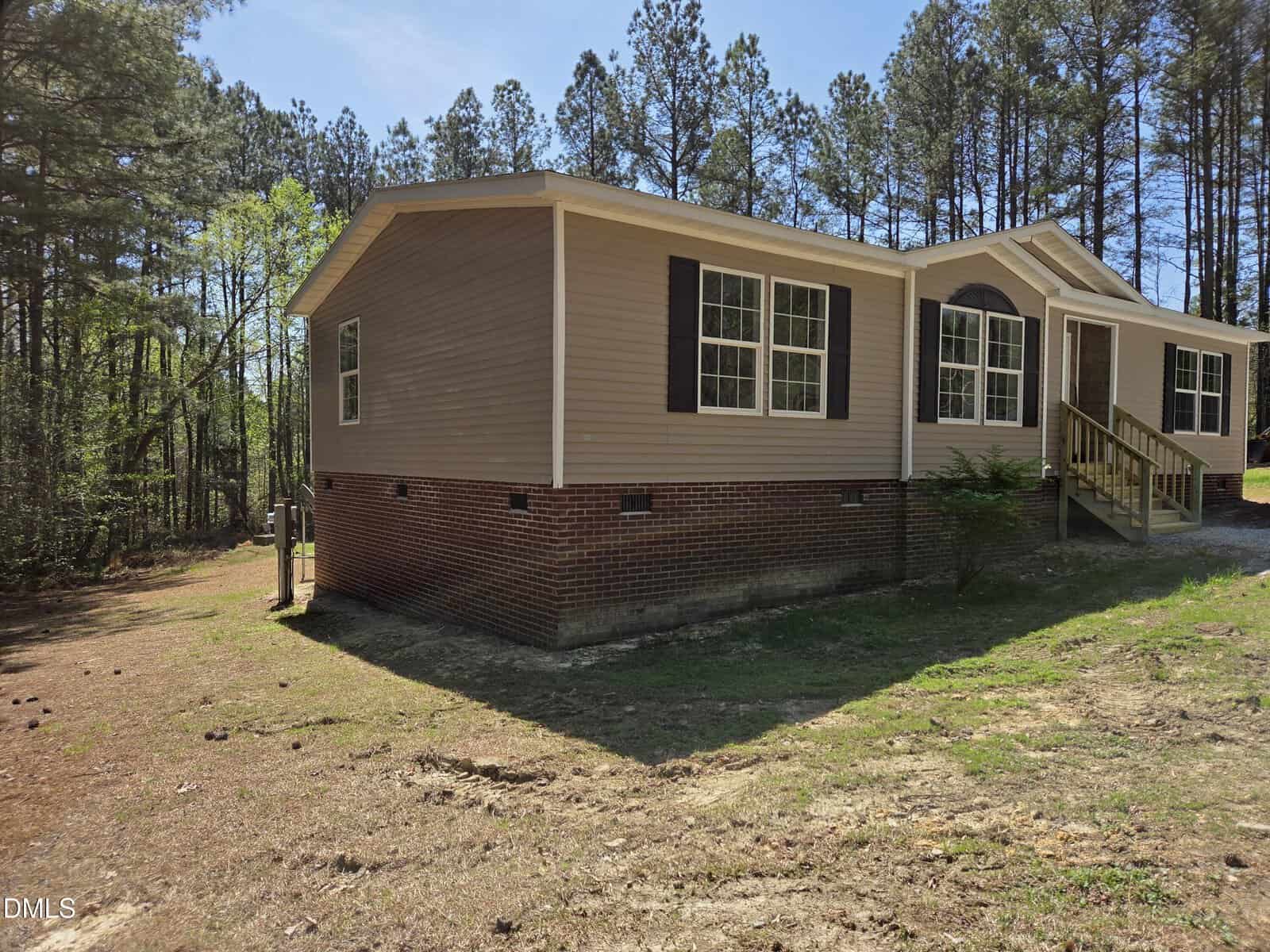 A tan manufactured home with brick foundation sits on a dirt lot, surrounded by tall pine trees under a clear blue sky. The house features white trim, dark shutters, and a small wooden porch with steps.