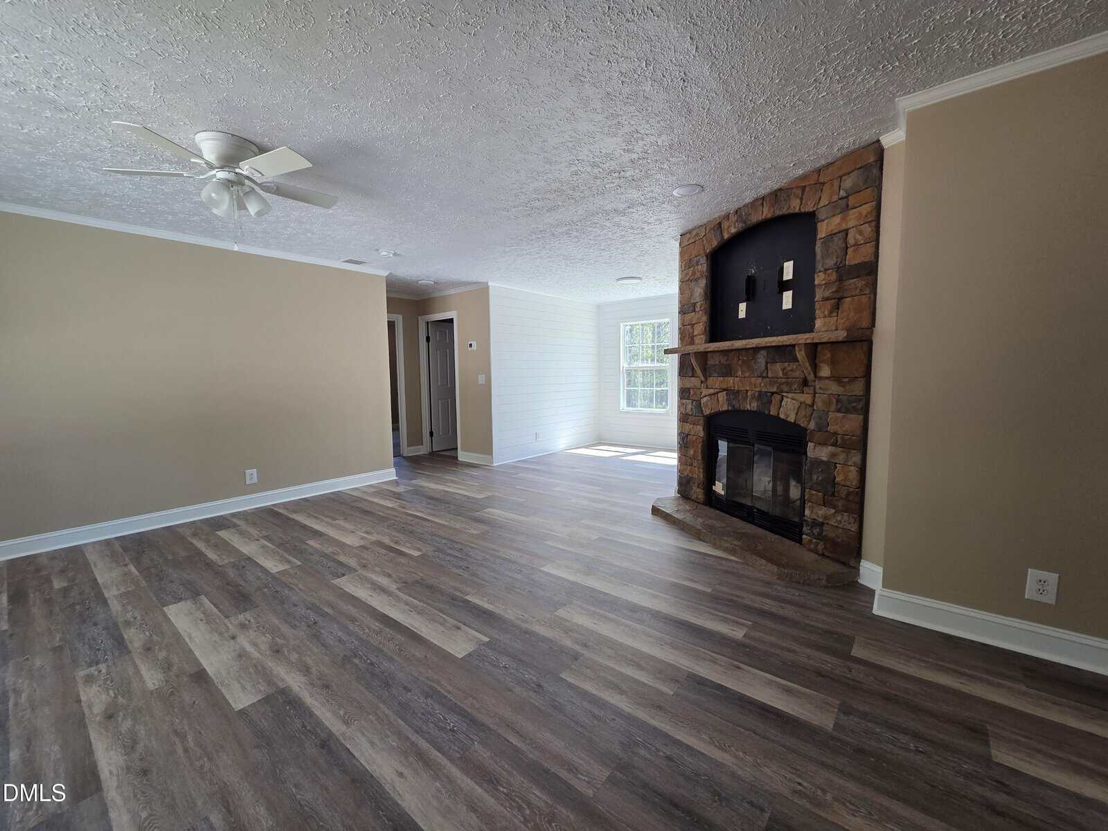 Spacious living room with wood-pattern vinyl flooring, beige walls, a white ceiling fan, and a stone fireplace. Sunlight streams in from windows near the dining area.
