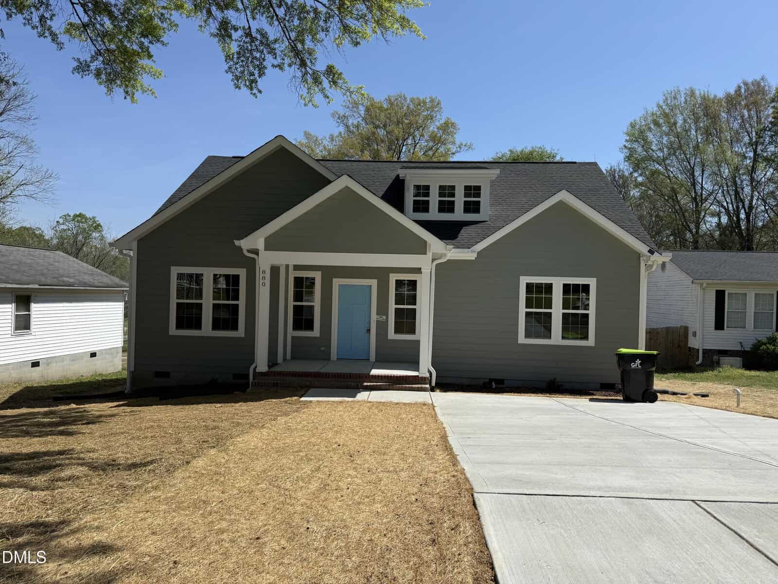 A single-story house with gray siding, white trim, and a light blue front door, featuring a small porch and a dormer window. The driveway is concrete, and the lawn is patchy with dry grass.