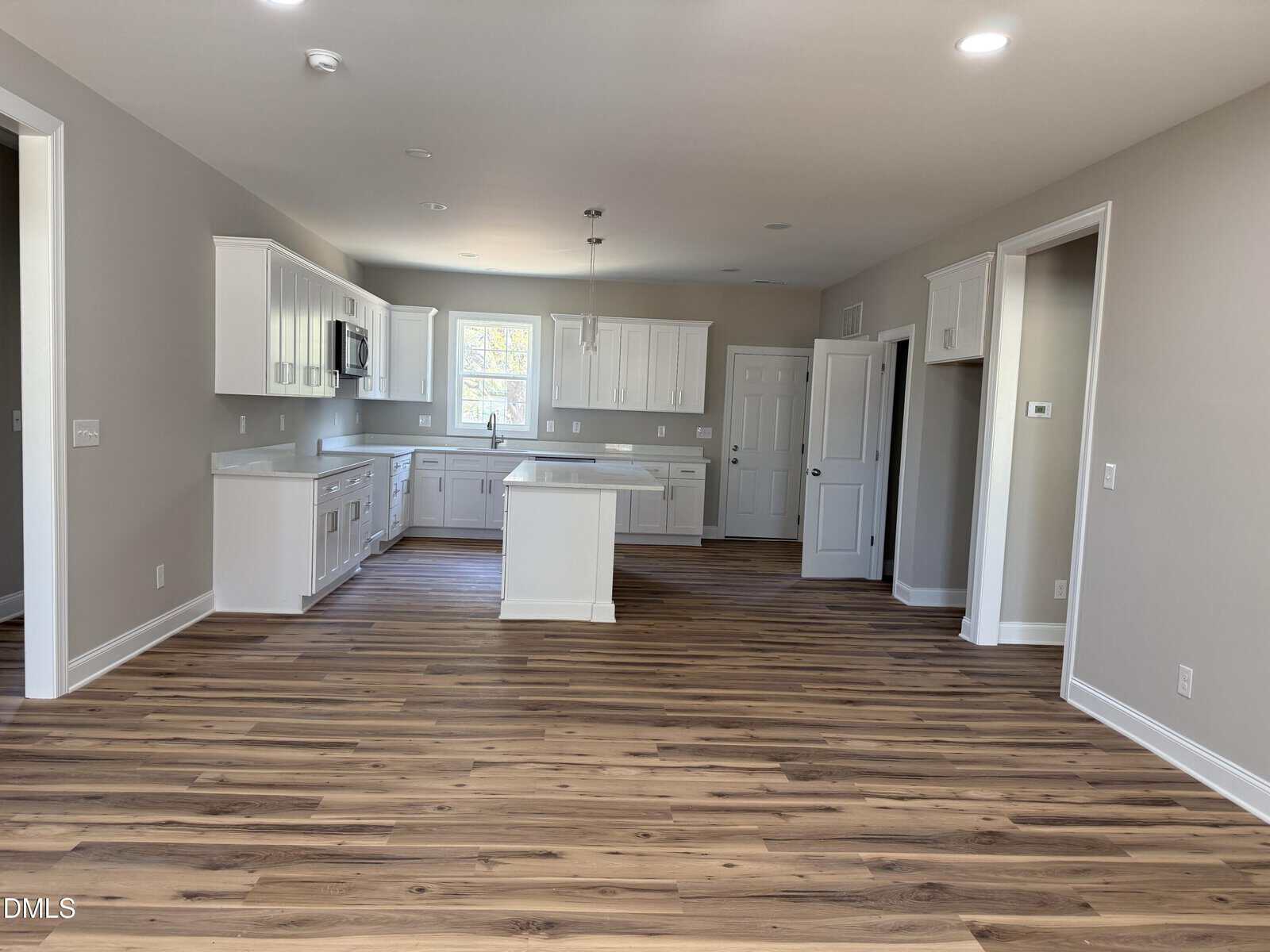 A modern kitchen with white cabinets, a center island, and stainless steel fixtures. The room has wood-style flooring, neutral walls, and recessed lighting. There are no appliances or furniture present.
