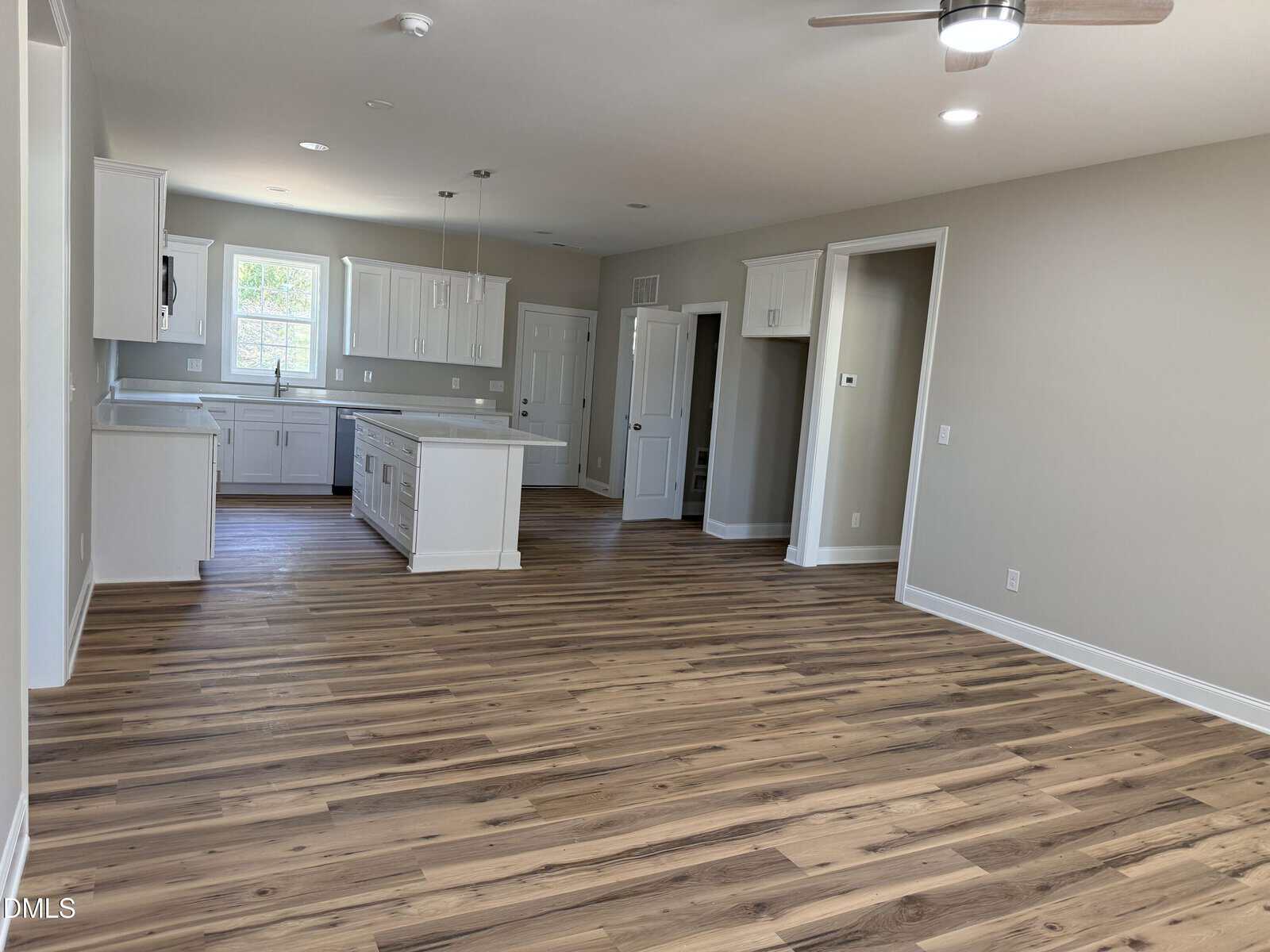 Open-plan kitchen and living area with light wood flooring, white cabinets, an island, and recessed lighting. The walls are light beige and a ceiling fan is visible. There are several windows and a door to the outside.
