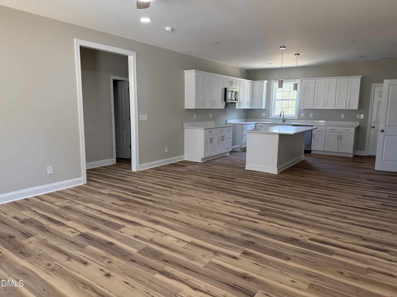 Open concept kitchen and living area with light wood floors, white cabinets, granite countertops, and stainless steel appliances. The room is empty, featuring neutral walls and natural light from multiple windows.
