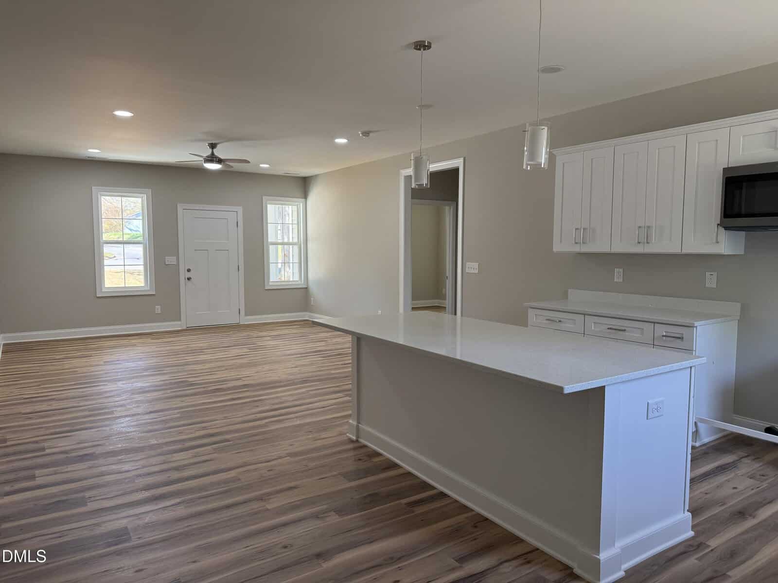 Open-concept kitchen and living area with light wood flooring, white cabinets, a large kitchen island, pendant lights, and neutral-colored walls. There are windows and a door along one wall, letting in natural light.