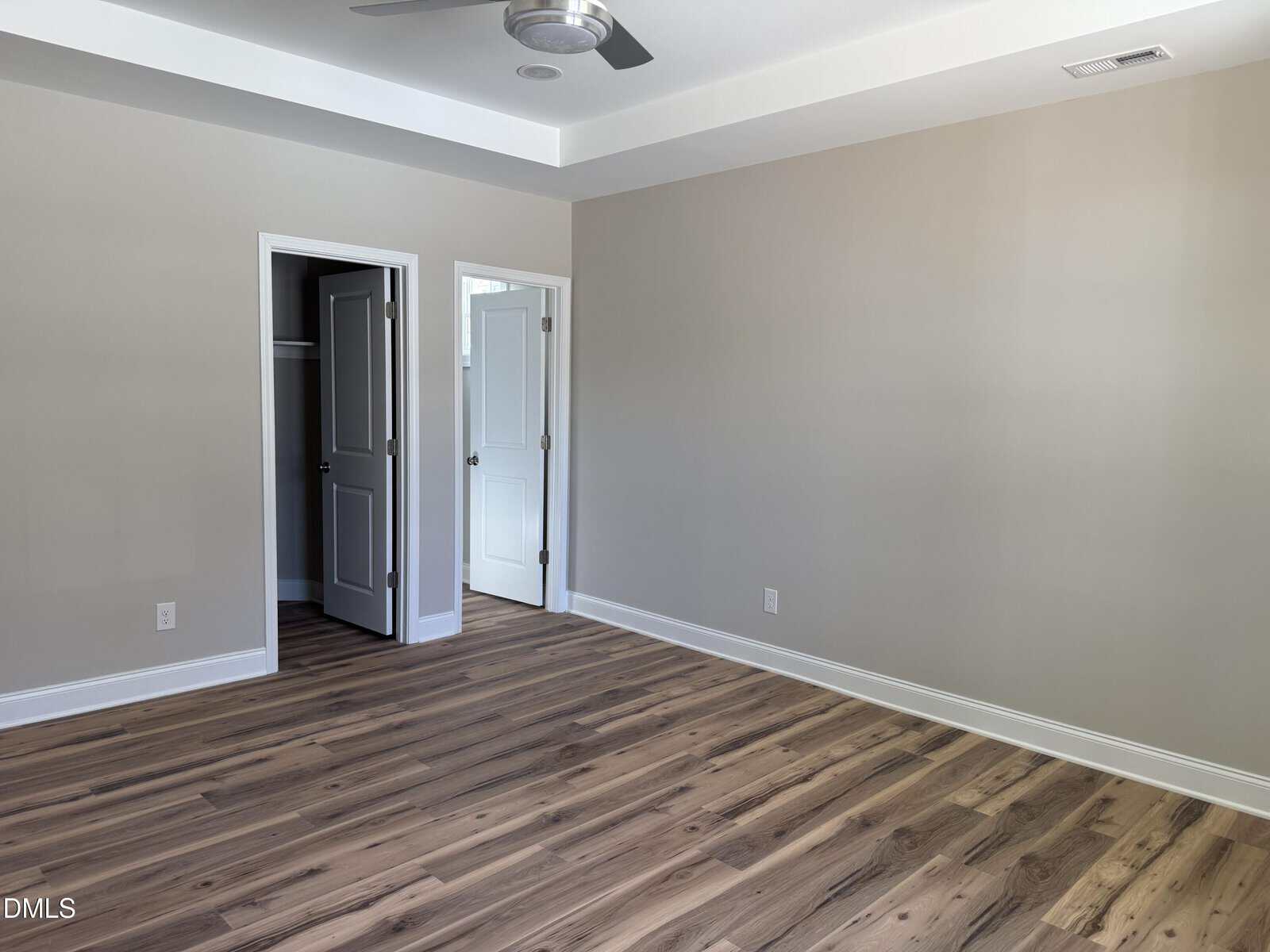 Empty room with light beige walls, wood-style flooring, a ceiling fan, and two open doors. One door leads to a closet, while the other opens to a hallway with light streaming in.
