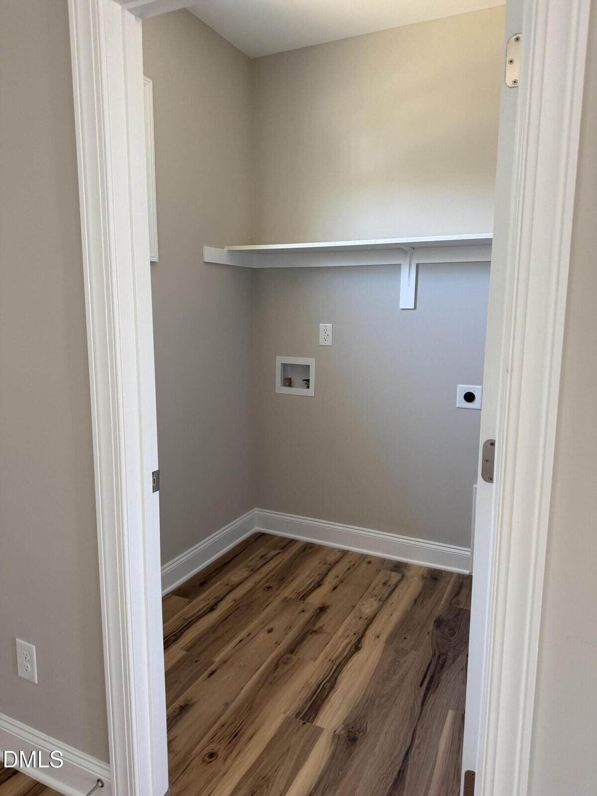 A small empty laundry room with light-colored walls, wood flooring, a white shelf, and hookups for a washer and dryer on the back wall.