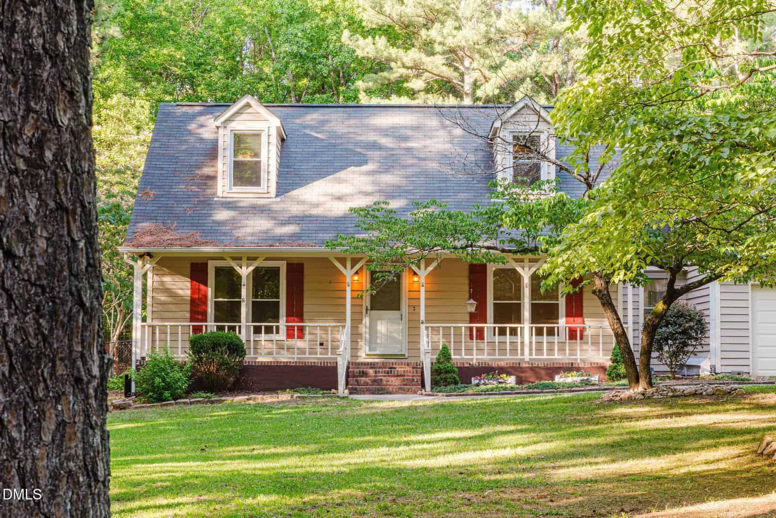 A beige, two-story house with a front porch, red shutters, and a sloped roof sits amid green trees and a grassy yard at 709 Appaloosa Way, Wake Forest, on a sunny day.