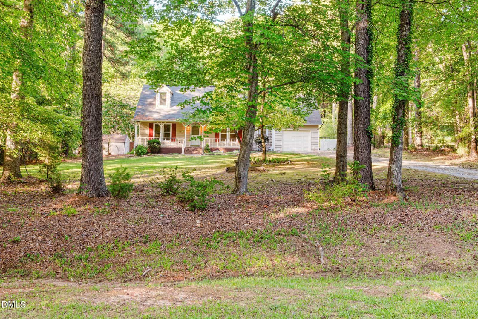 A house with a large front porch and attached garage is nestled among tall trees, surrounded by a spacious, shaded yard with patches of grass and scattered shrubs. A gravel driveway curves toward the house.