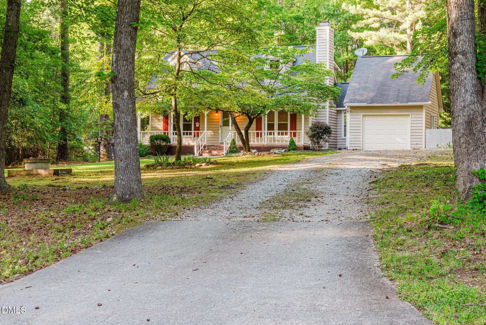 A light-colored house with a front porch, red shutters, and a single-car garage sits among tall trees. A gravel driveway leads to the house, surrounded by a green, wooded yard.